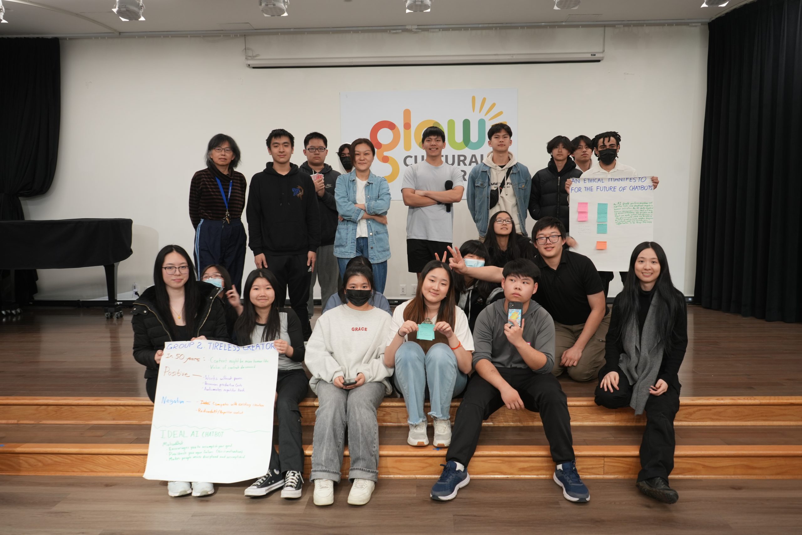 Group photo of "Whose AI?" workshop participants in Glow Cultural Center's ballroom - a row of highschool students sit on the stage steps while others stand behind them holding a colorful worksheet of the "Ethical AI Manifesto" created. 
